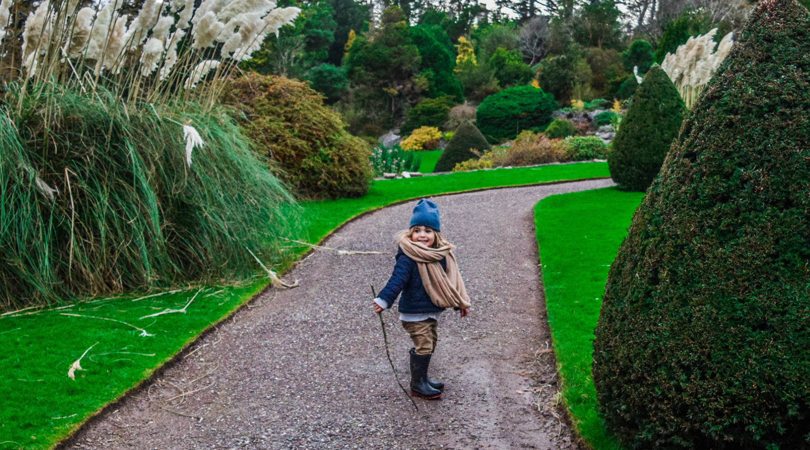 little girl with winter clothes on including a blue hat and scarf holds a walking stick while on a path dotted with green bushes, grass and greenery
