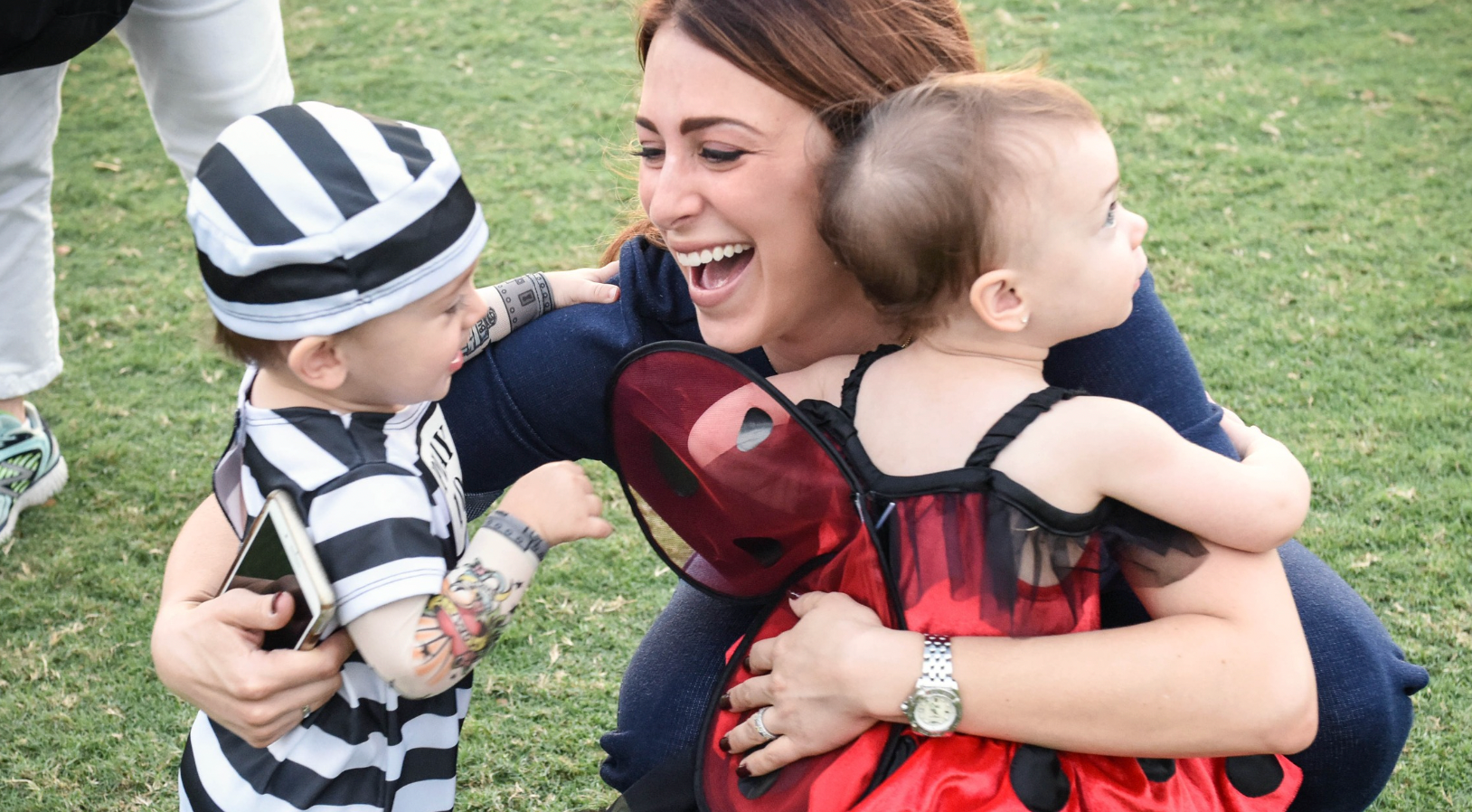 very happy smiling woman hugging two toddlers in halloween costumes, one is a little prisoner and the other a ladybug