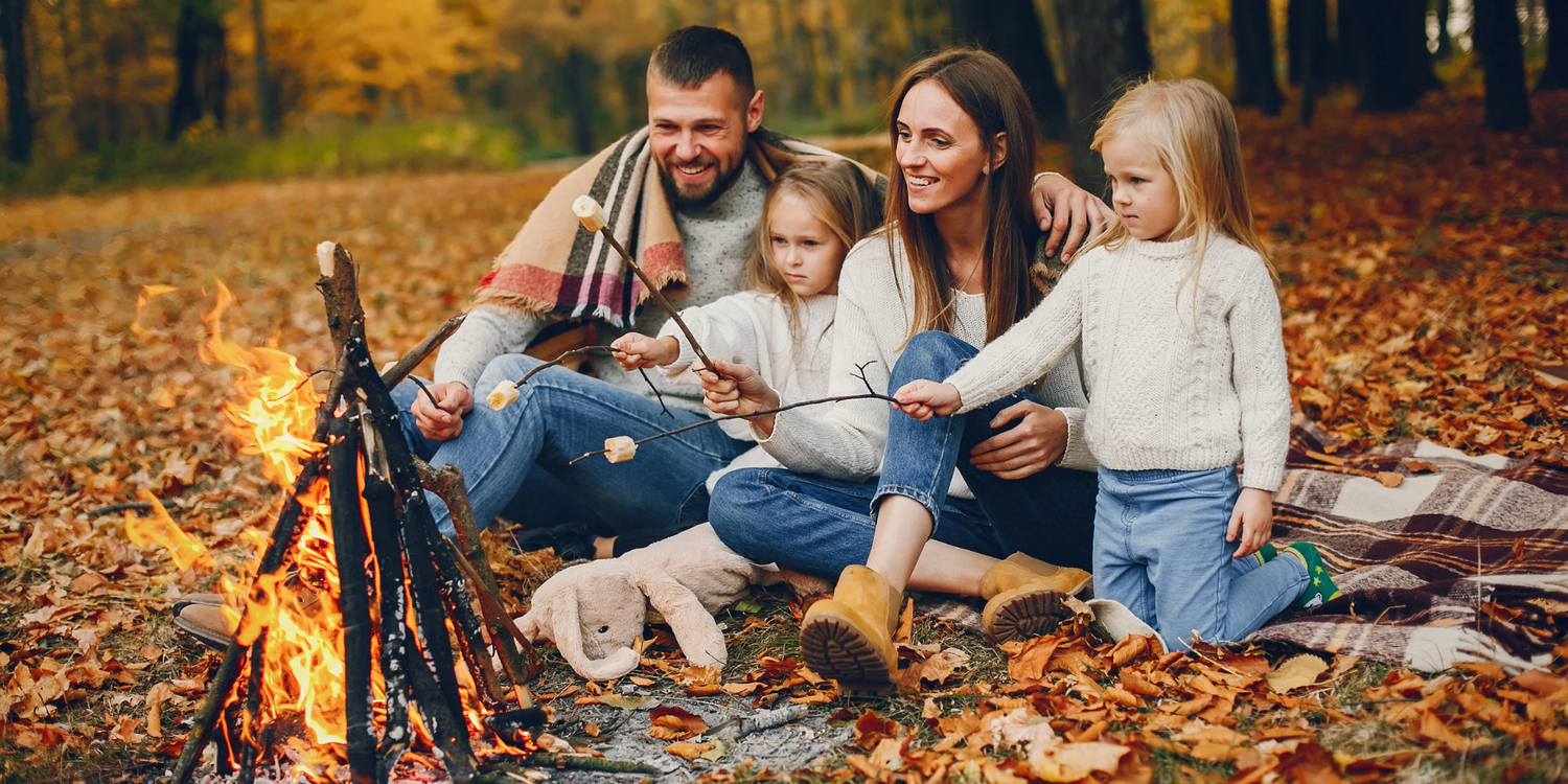 Family of four with mom, dad and two little girls roasting marshmallows at a campfire surrounded by fall leaves in orange and yellow