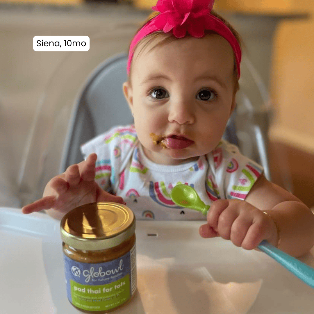 baby with food around their mouth in high chair holding a spoon reaching for jar of Globowl Pad Thai for Tots