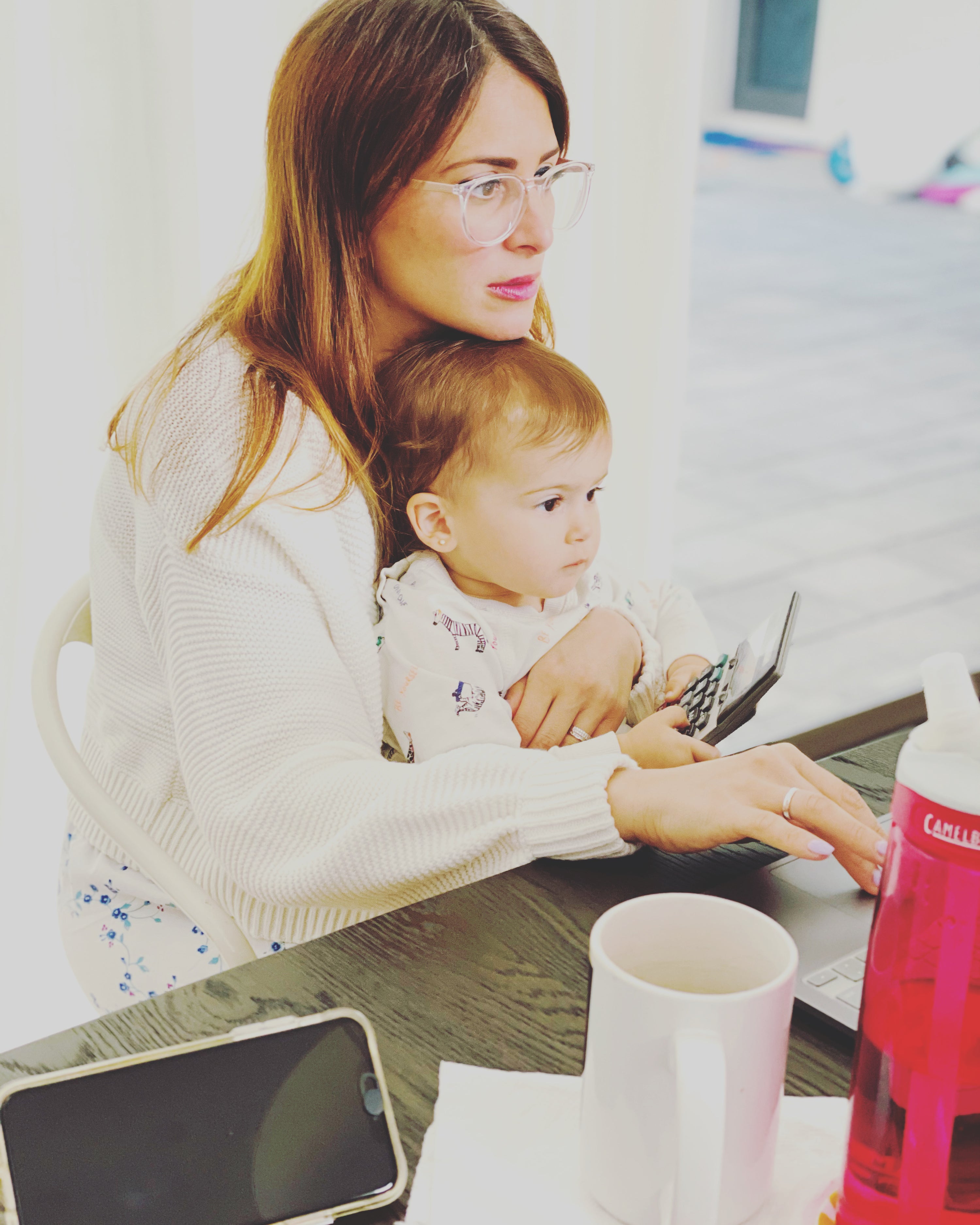 Woman in glasses and a white sweater holding her baby while working on the computer