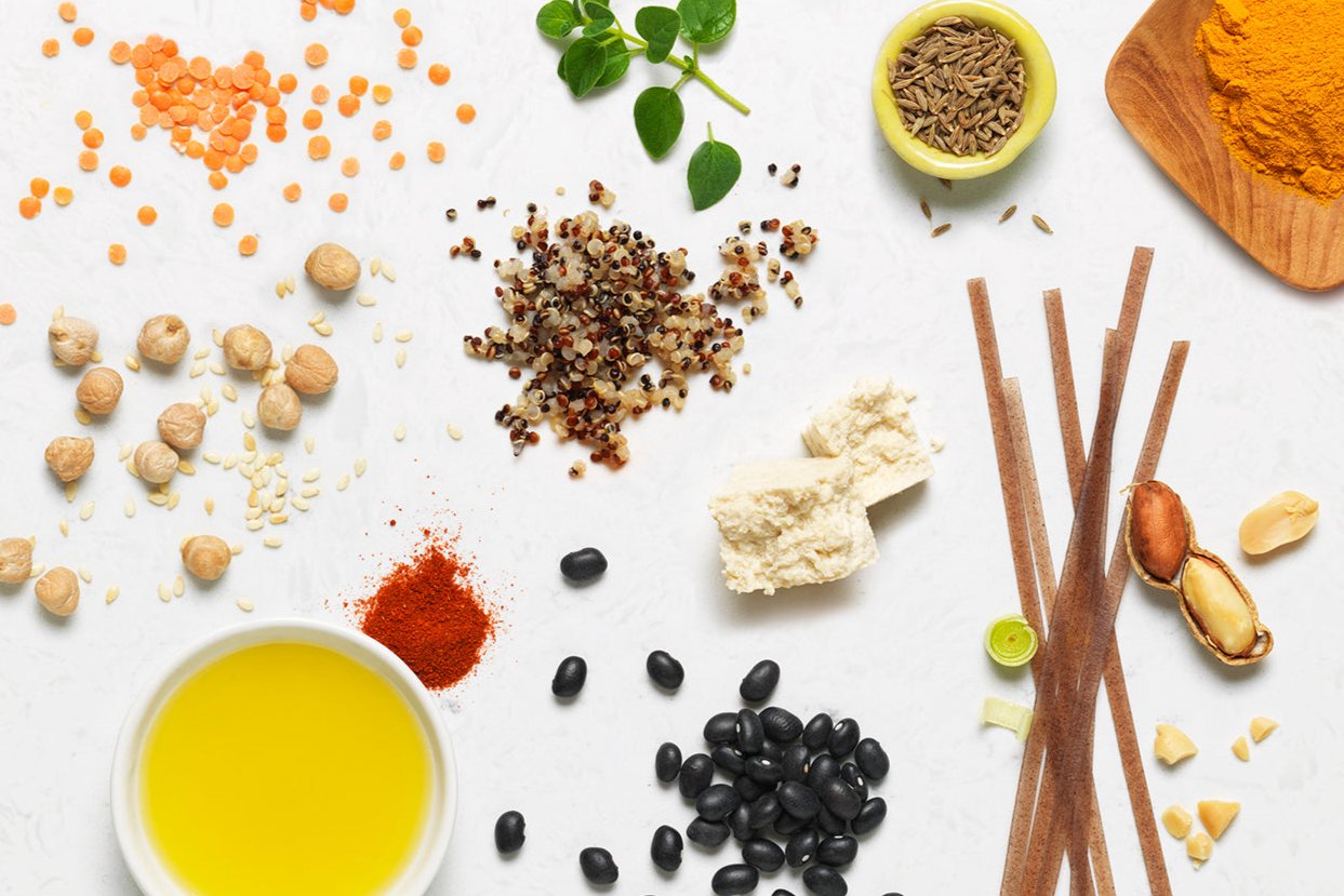 ingredients (red lentils, chickpeas, sesame seeds, quinoa, tofu, dry rice noodles, peanuts, black beans and spices) are scattered in loose piles on a white marble surface