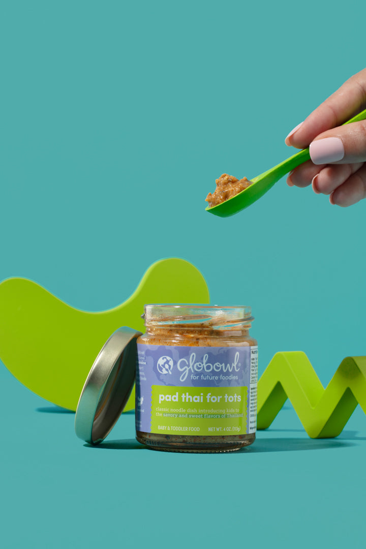 open jar of Pad Thai for Tots on a teal background with a hand above holding a small spoon of food
